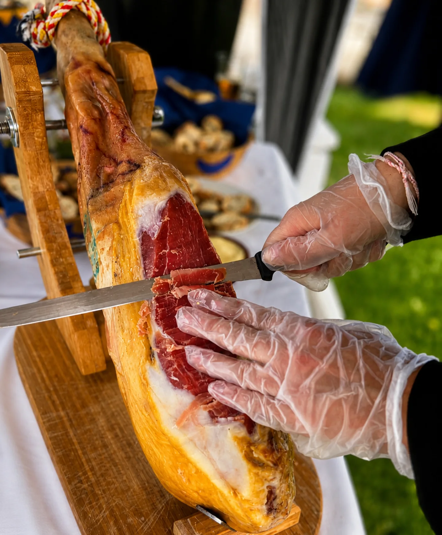 Découpe à la main d'un jambon cru à l'os sur son support en bois, lors d'une animation traiteur en extérieur avec gants de protection.