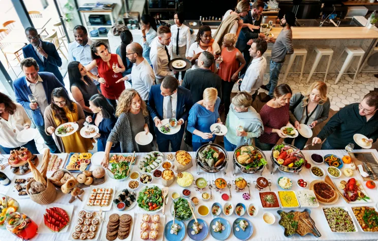 Vue plongeante d'une grande tablée conviviale lors d'un événement traiteur avec buffet abondant et invités réunis autour des assiettes colorées.