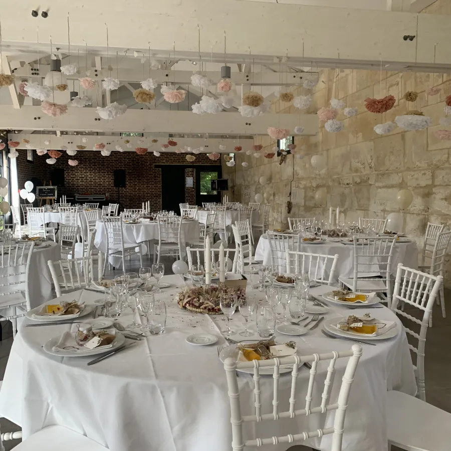 Salle de réception aux murs en pierre avec tables rondes nappées de blanc, chaises Chiavari et centres de table végétaux pour un mariage.