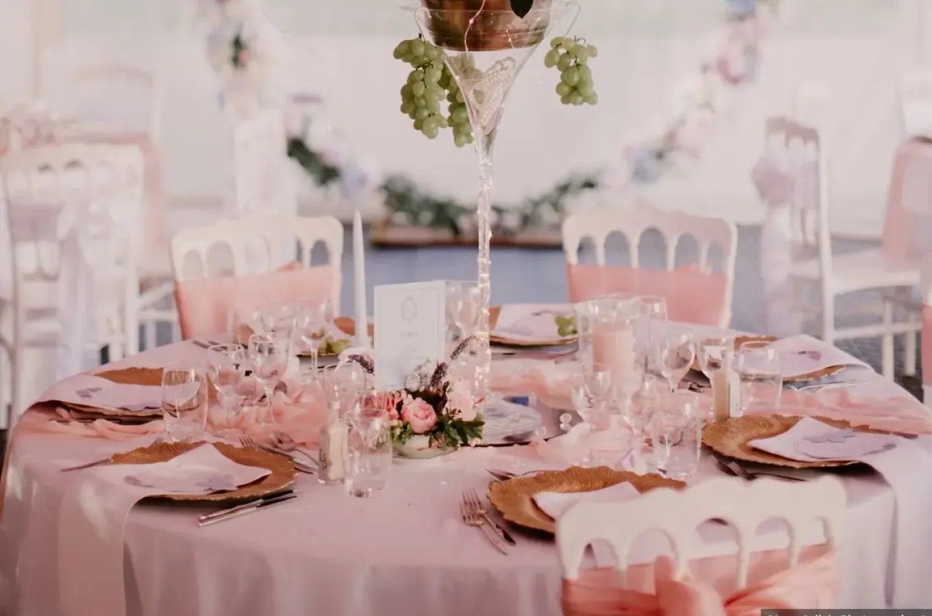 Table ronde de mariage dressée en rose poudré avec assiettes dorées, verres martini garnis de fleurs et grand centre de table fleuri sous chapiteau.