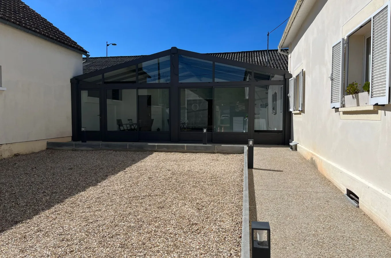 Veranda contemporaine a structure noire et toit vitre attenante a une maison, avec cour en gravier, espace evenementiel exterieur disponible a la location.