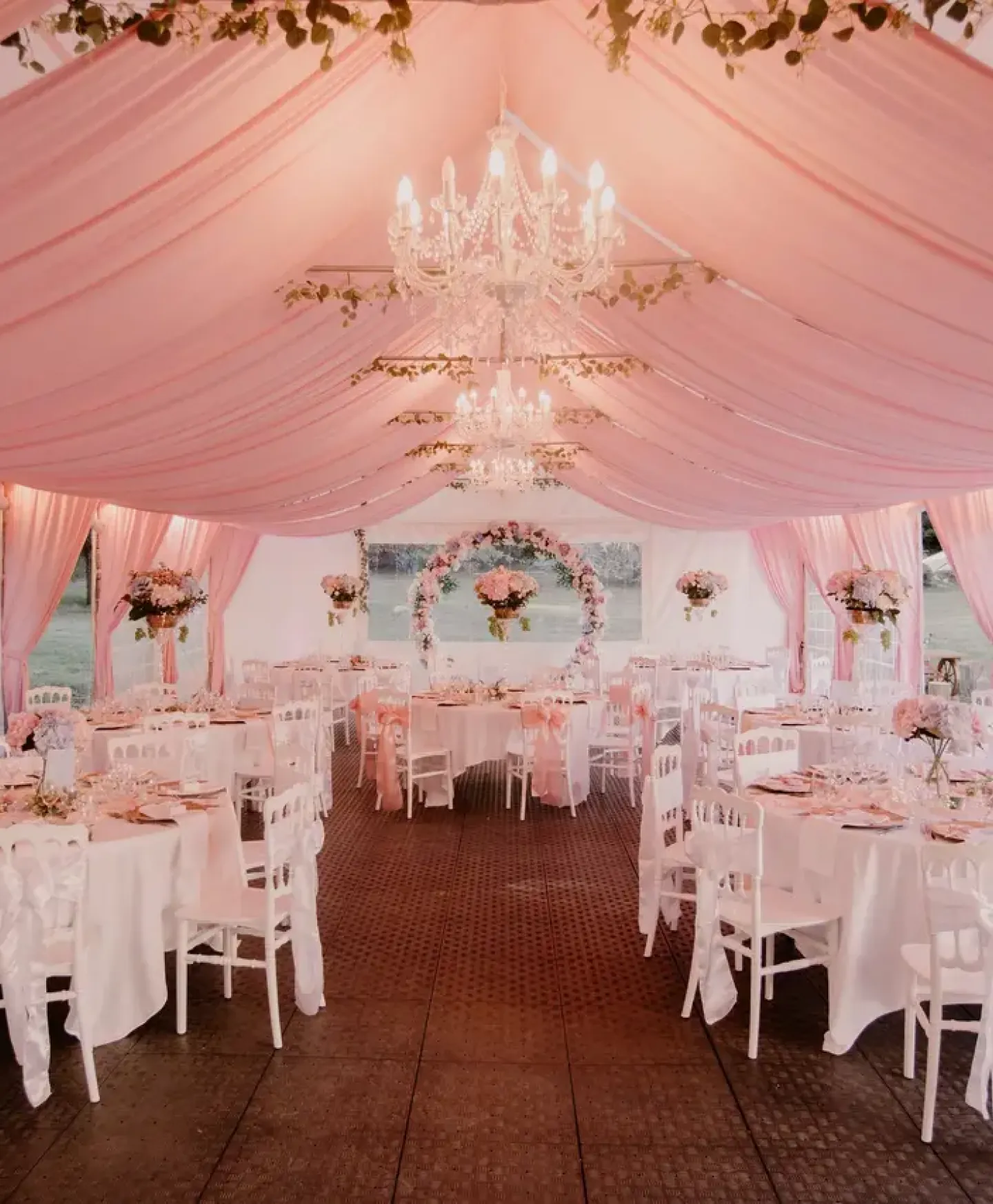 Salle de reception de mariage sous tonnelle drapee de tissu rose pale, avec lustres en cristal, tables rondes nappees de blanc et arche fleurie pour une ceremonie romantique en exterieur.