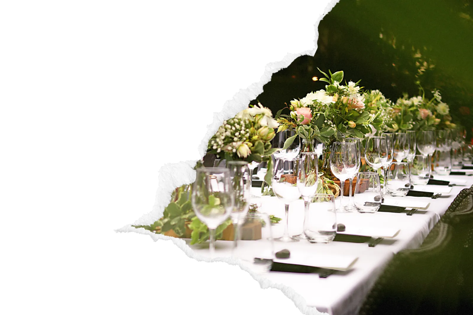Table de reception de mariage elegamment dressee avec nappe blanche, alignement de verres a pied en cristal, centres de table fleuris de roses et verdure, couverts sur sets sombres pour un service traiteur haut de gamme dans la zone de Rouen. Image avec bord dechire decoratif sur fond noir.