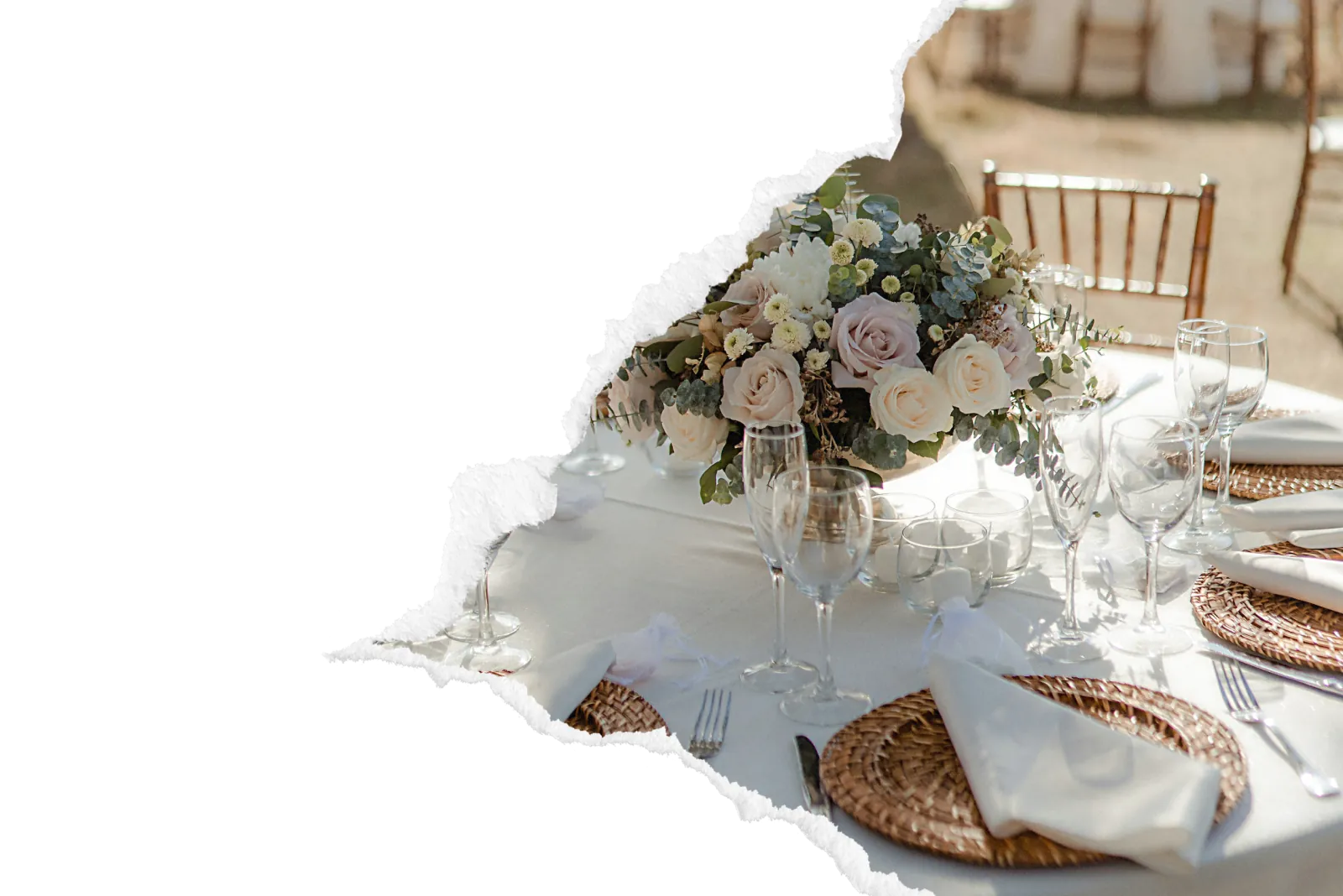 Table de mariage dressee en exterieur avec bouquet de roses pastel creme et rosees, verres en cristal, sous-assiettes en osier tresse et nappage blanc immaculé pour une reception elegante par le traiteur A Votre Gout dans la zone de Gisors. Image avec bord dechire decoratif sur fond noir.