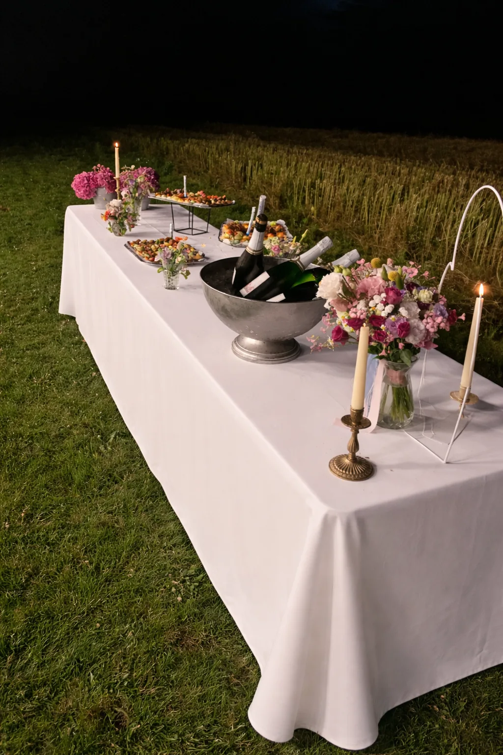Buffet de mariage dressé en plein champ à la tombée de la nuit, nappe blanche immaculée, seau à champagne, bouquets pastel et bougies allumées composent une atmosphère champêtre raffinée.
