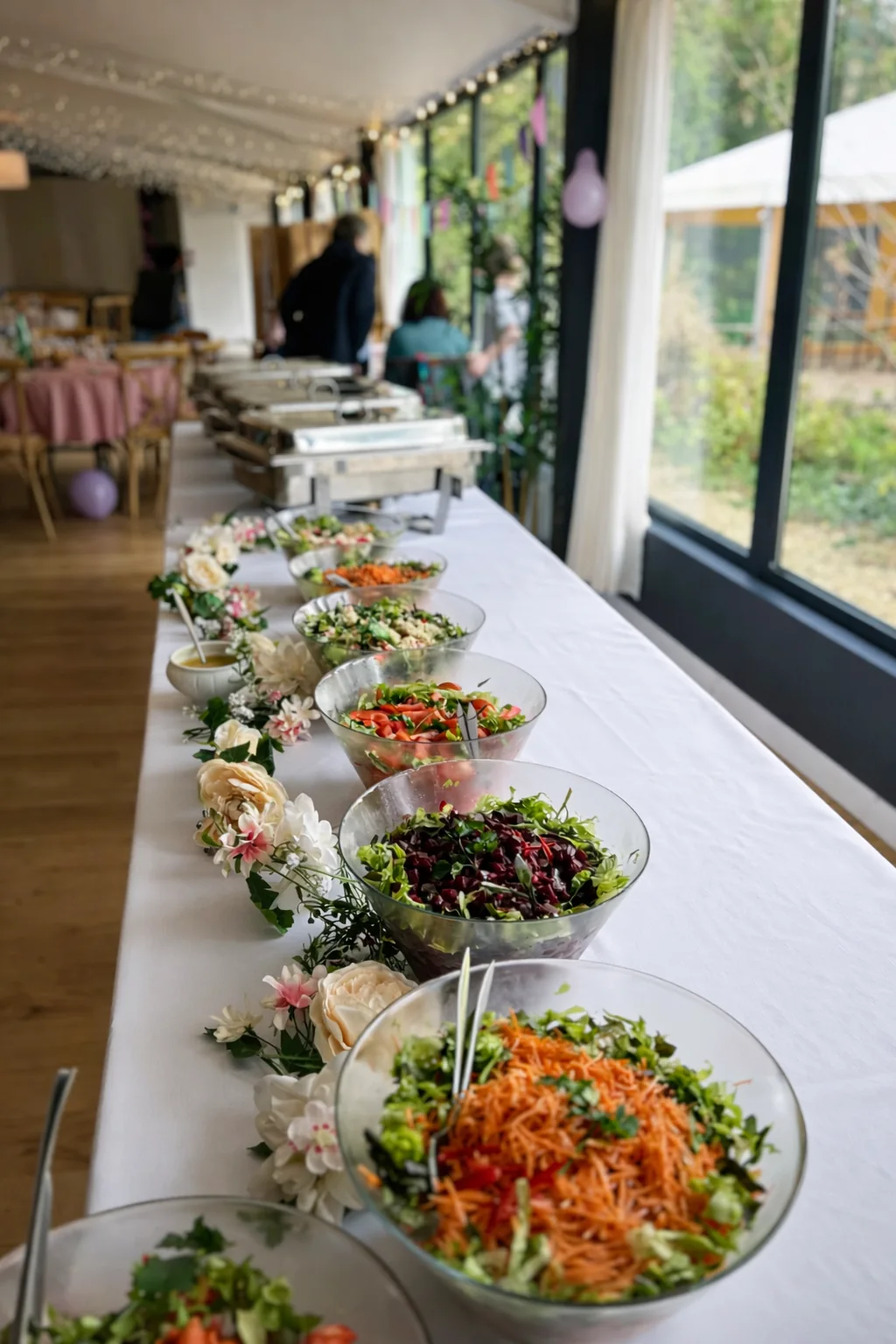 Long buffet de salades composées colorées dressé en saladiers transparents sur nappe blanche, décoré de fleurs et installé dans une salle de réception lumineuse.