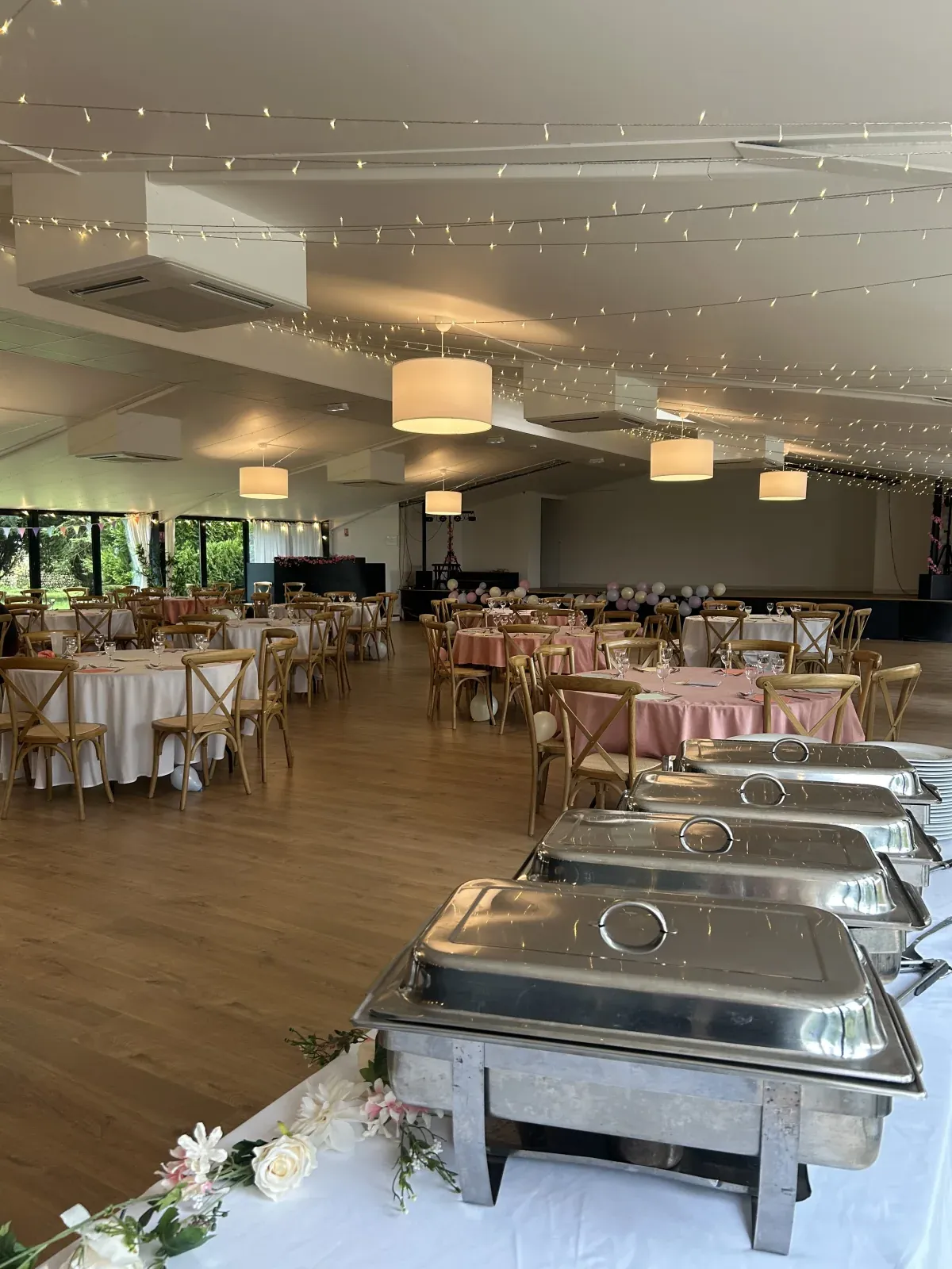 Salle de réception élégante avec buffet chaud installé sur chafing-dishes en inox, tables habillées de nappes roses et guirlandes lumineuses au plafond.