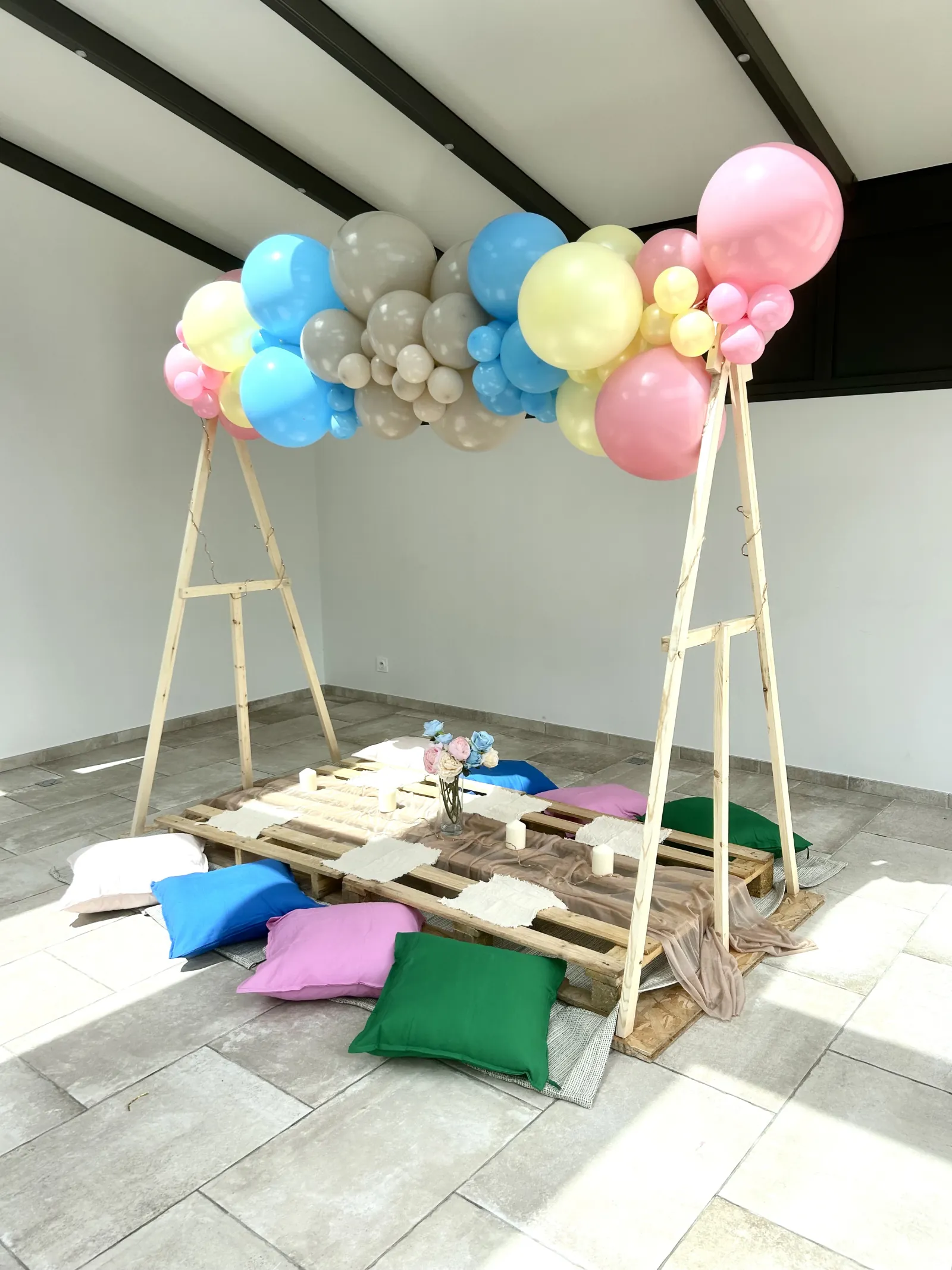 Espace de réception décoré d'une arche de ballons pastel et d'une palette en bois entourée de coussins colorés, prêt pour un événement convivial.