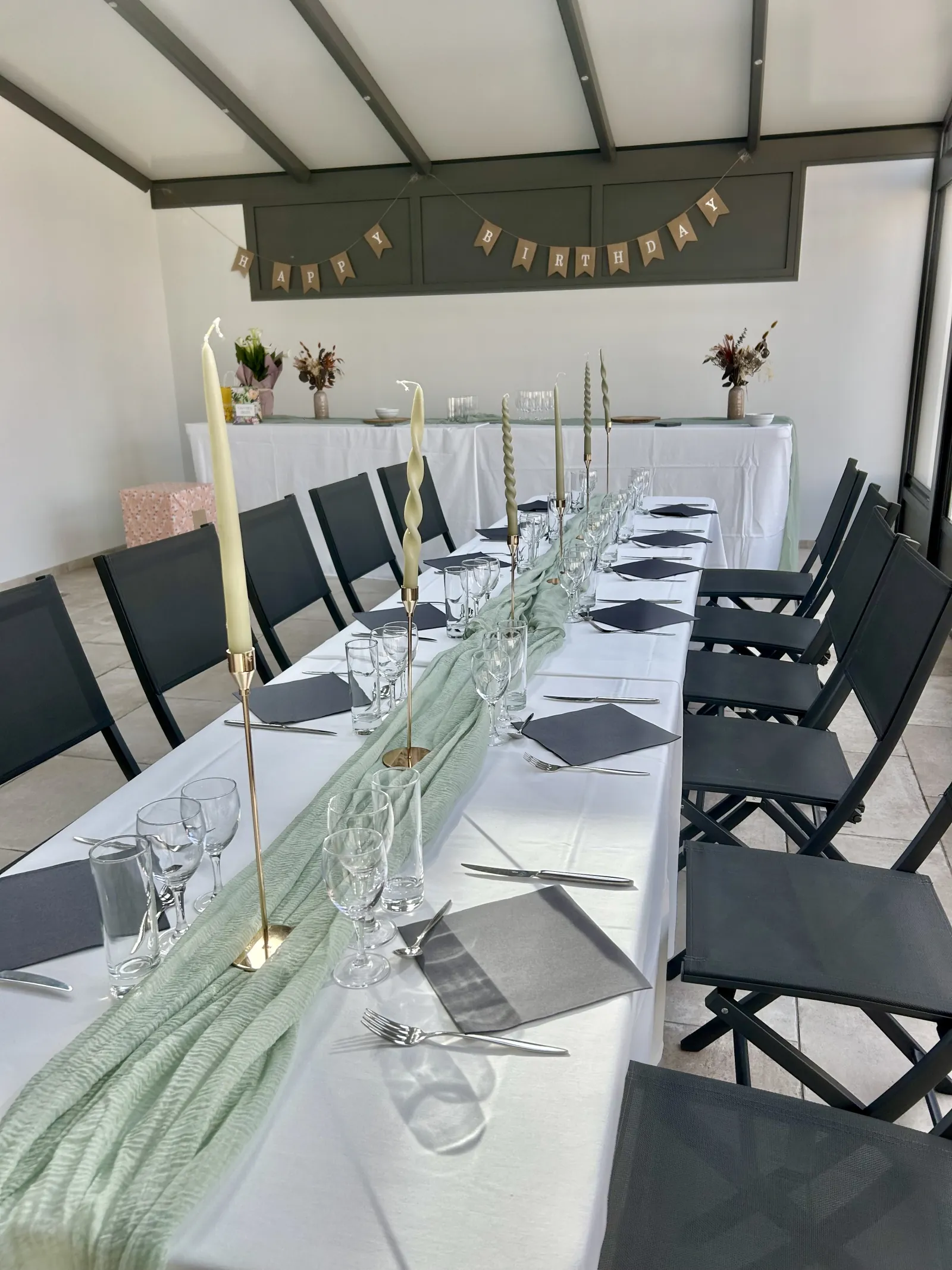Longue tablée dressée pour un anniversaire avec chemin de table vert d'eau, bougies hautes et guirlande Happy Birthday dans une salle lumineuse.