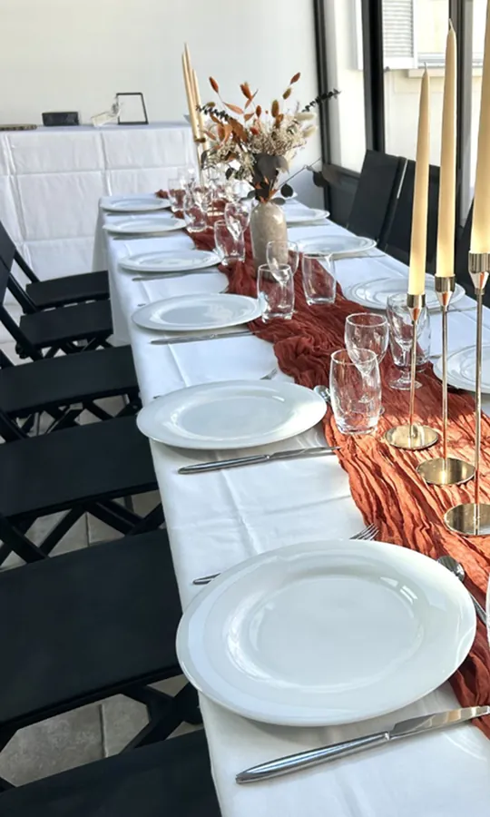 Table d'événement dressée avec assiettes blanches, chemin de table terracotta et chaises noires alignées dans une salle moderne.