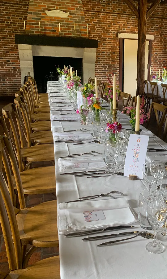 Longue table de banquet dressée avec nappes blanches, vaisselle et bougies dans une salle de réception en briques.