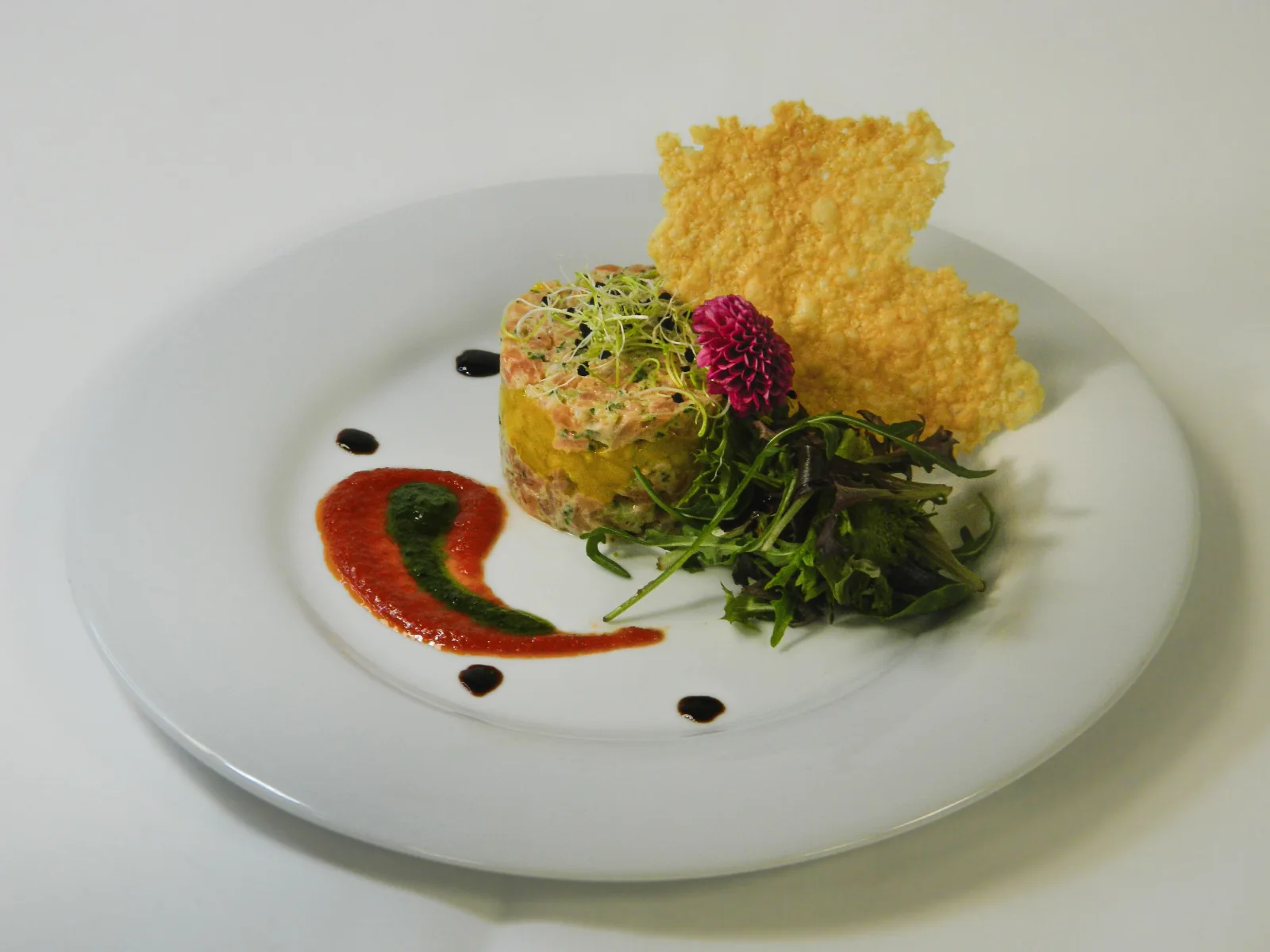Tartare de saumon dressé en cercle avec tuile de parmesan croustillante, mesclun, fleur comestible et coulis bicolore tomate-basilic.