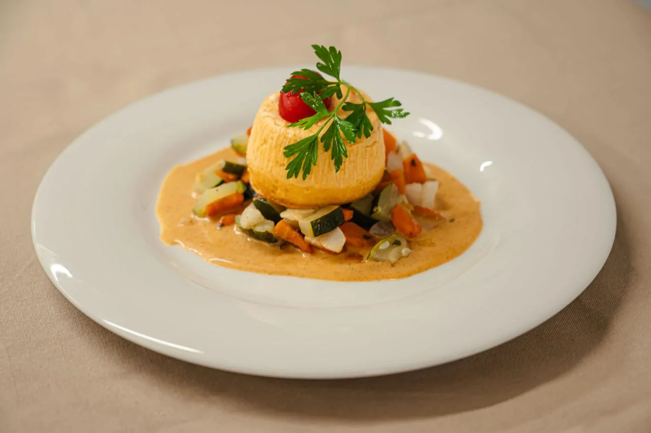 Flan moelleux dressé sur une brunoise de légumes nappée d'une sauce corail, finition persil plat et tomate cerise sur assiette blanche.