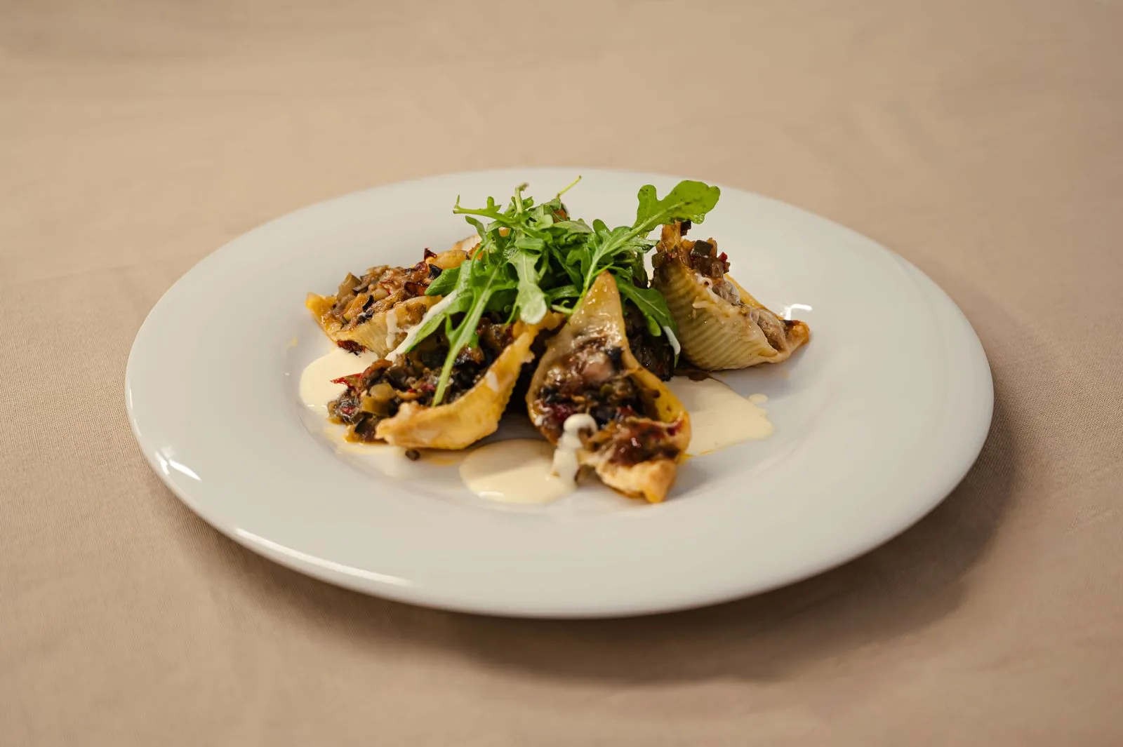 Conchiglioni farcis aux légumes confits et tomates séchées, bouquet de roquette fraîche et crème onctueuse dressés sur assiette blanche.