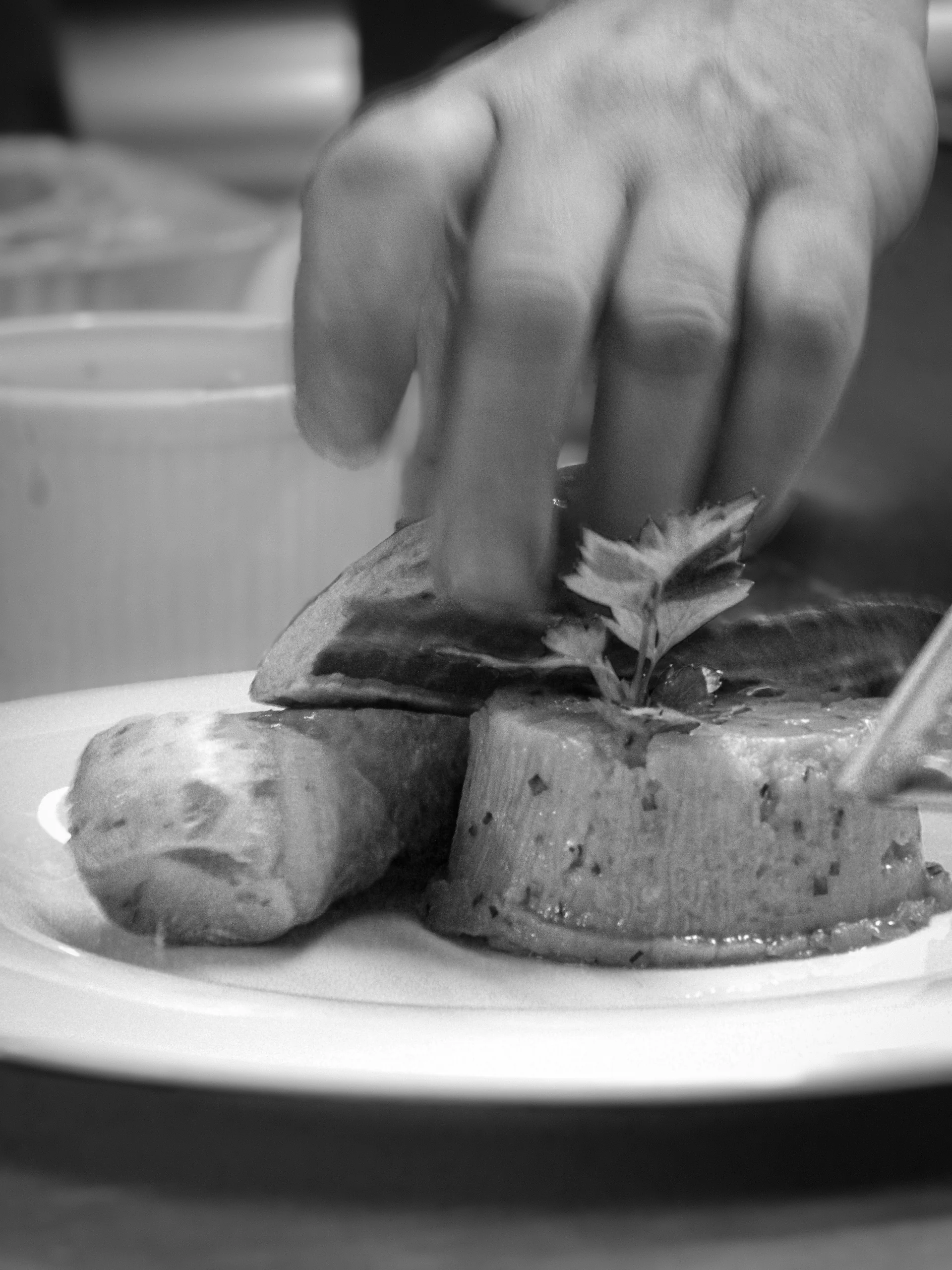 Main du chef qui dépose délicatement une herbe aromatique sur une terrine, image en noir et blanc qui dit la minutie du dressage maison.