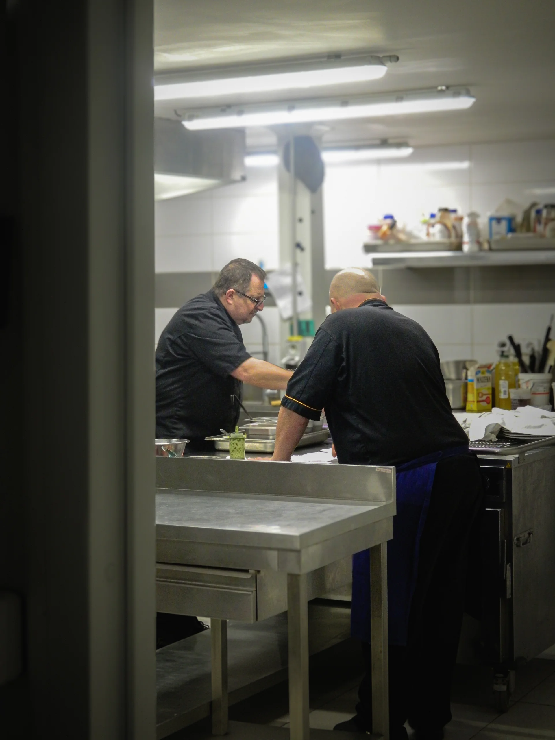 Deux membres de l'équipe À Votre Goût en plein service dans le laboratoire de cuisine, concentrés autour du plan de travail inox — les coulisses du traiteur.