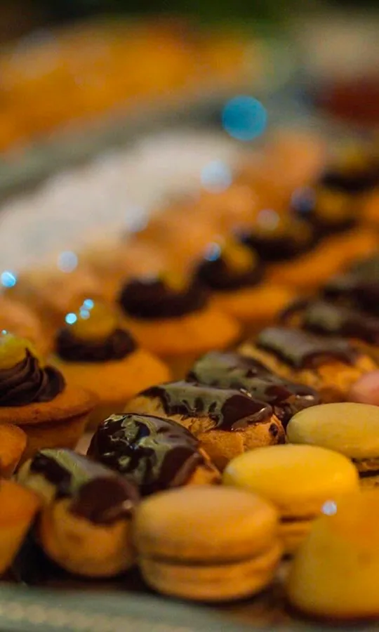 Buffet de mignardises sucrées en gros plan : éclairs au chocolat, macarons jaunes et petits choux glacés disposés en éventail dans une ambiance lumineuse.