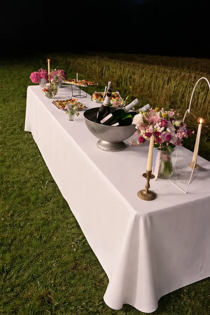 Buffet de mariage dressé en plein champ à la tombée de la nuit, nappe blanche immaculée, seau à champagne, bouquets pastel et bougies allumées composent une atmosphère champêtre raffinée.