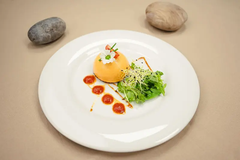 Plat raffiné dressé à l'assiette : dôme orangé surmonté d'une fleur comestible, mesclun de roquette et points de coulis sur fond beige.