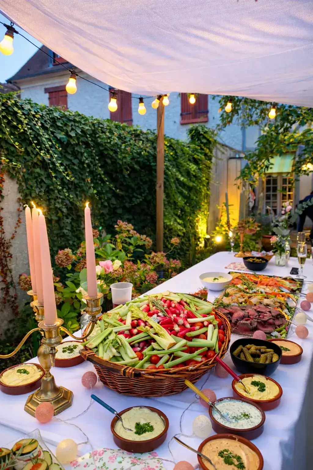 Buffet apéritif présenté sur longue table en jardin extérieur sous tonnelle illuminée de guirlandes lumineuses, avec panier de crudités, houmous, bougies et fleurs séchées en ambiance conviviale nocturne.