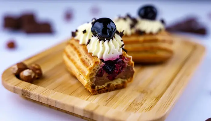 Bouchées gourmandes type mini-gaufres garnies de crème vanille, myrtilles et copeaux de chocolat, présentées sur planche en bambou avec noisettes.