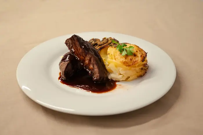Pavé de bœuf nappé d'une sauce au vin rouge, accompagné d'un gratin dauphinois doré et de champignons persillés sur assiette blanche.