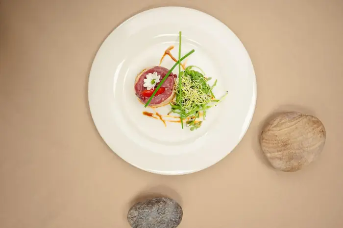 Tartare de bœuf en cercle, fleur de pâquerette et tomate cerise, pousses fraîches et ciboulette dressés en vue plongeante sur assiette blanche.