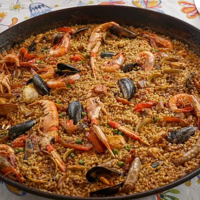Gros plan d'une paella genereuse aux langoustines, moules et crevettes servie dans une poele traditionnelle sur nappe a motifs floraux.
