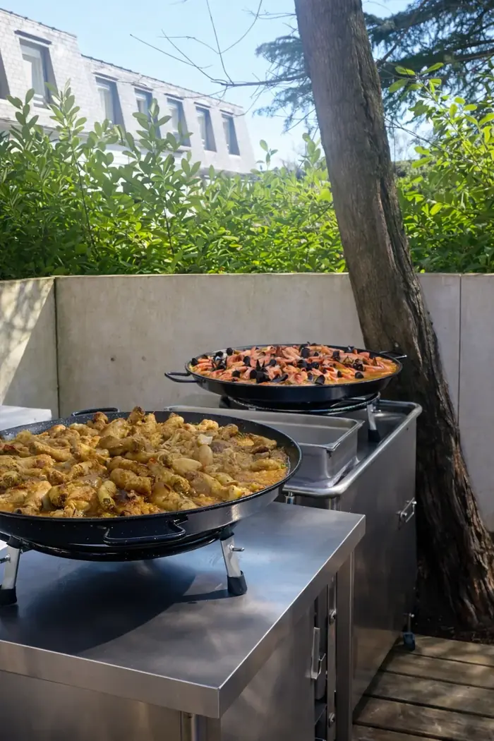 Duo de grandes paellas en cuisson sur dessertes inox en extérieur, l'une au poulet et l'autre aux fruits de mer, prêtes à être partagées lors d'un événement traiteur.