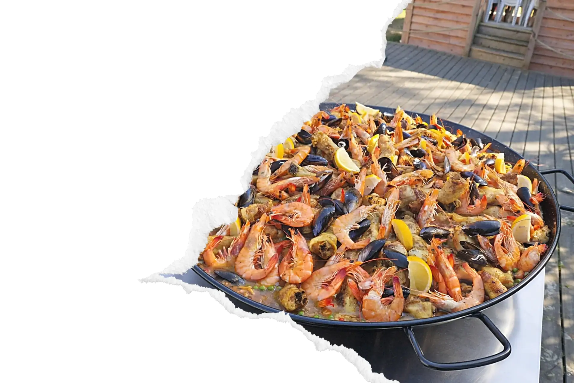 Paëlla géante aux fruits de mer (crevettes, moules, citron) préparée en grande poêle sur trépied, installée en extérieur sur terrasse en bois pour un événement convivial.