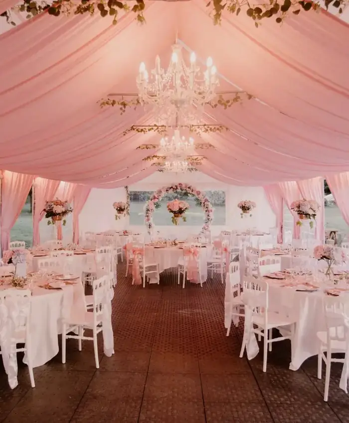 Salle de reception de mariage sous tonnelle drapee de tissu rose pale, avec lustres en cristal, tables rondes nappees de blanc et arche fleurie pour une ceremonie romantique en exterieur.