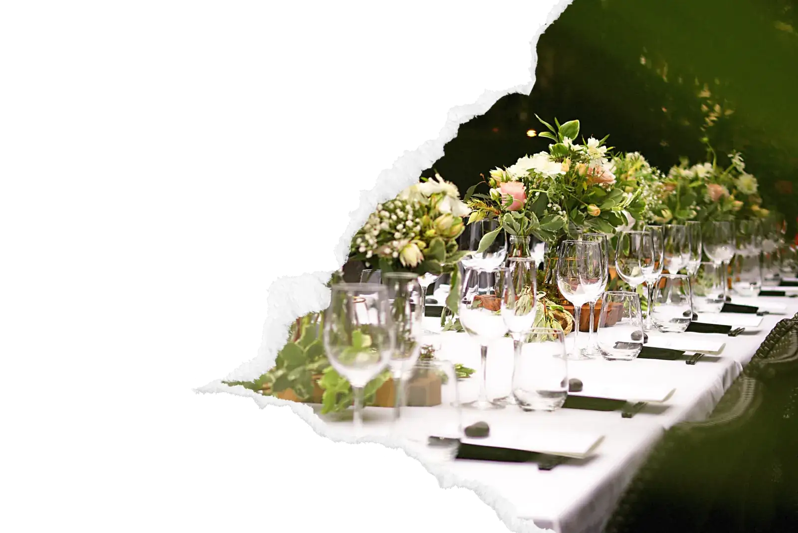 Table de reception de mariage elegamment dressee avec nappe blanche, alignement de verres a pied en cristal, centres de table fleuris de roses et verdure, couverts sur sets sombres pour un service traiteur haut de gamme dans la zone de Rouen. Image avec bord dechire decoratif sur fond noir.