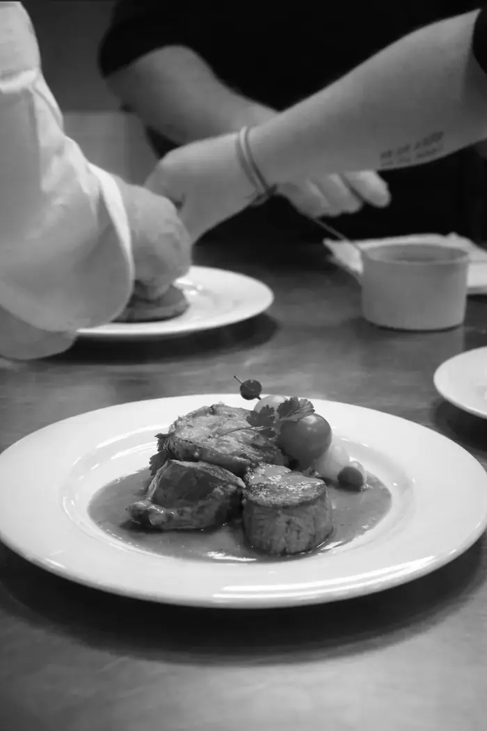 Le chef finalise le dressage d'une assiette de viande et garniture en cuisine, scène en noir et blanc qui capte le geste précis du métier.