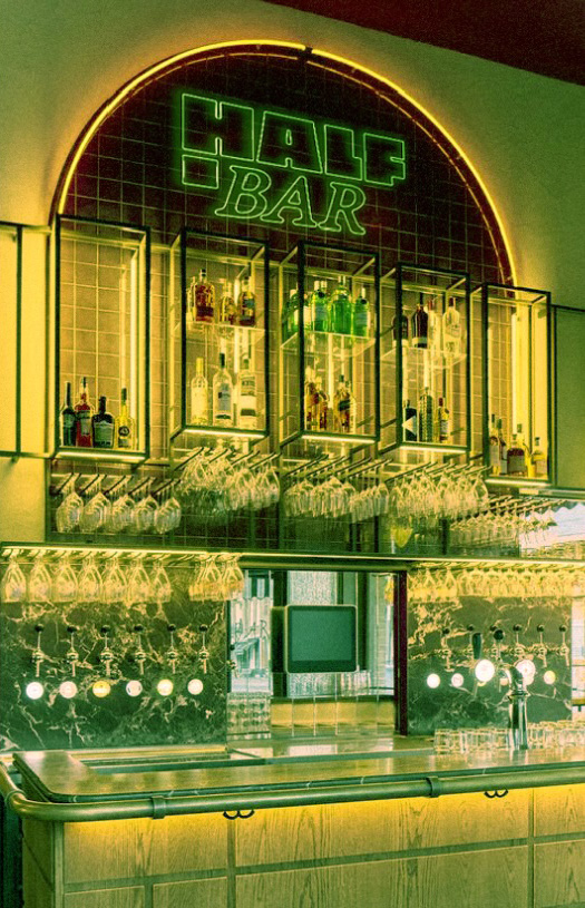 Intérieur de bar moderne avec un comptoir en bois éclairé, un mur de bouteilles et des verres suspendus, enseigne lumineuse verte "HALF BAR".