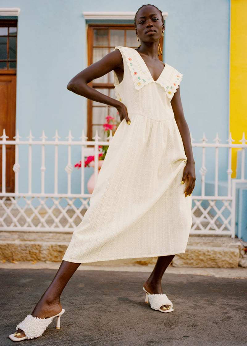 Femme noire portant une robe blanche mi-longue à col large avec broderies florales, posant devant une maison colorée avec une clôture blanche.