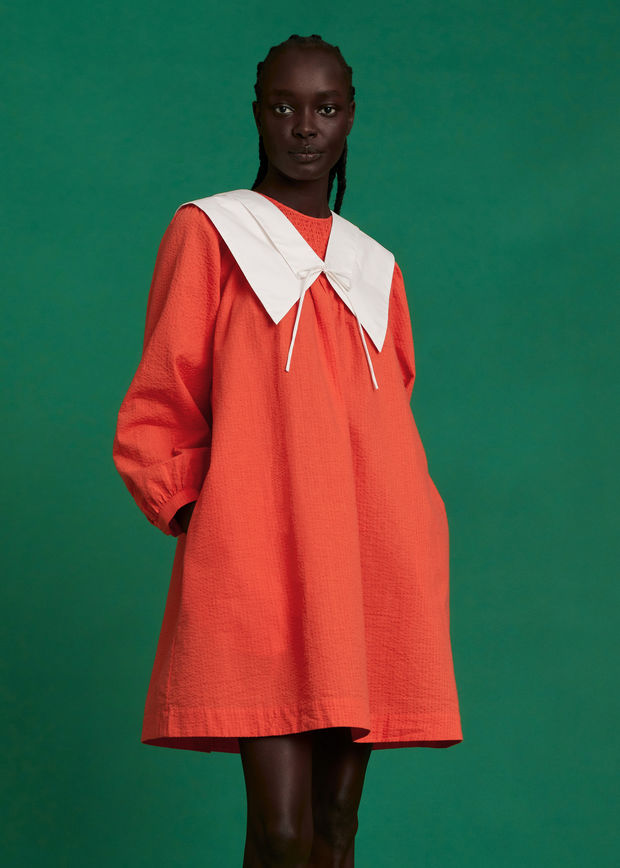Femme portant une robe ample orange à manches longues avec un grand col blanc à nouer, posant devant un fond vert.