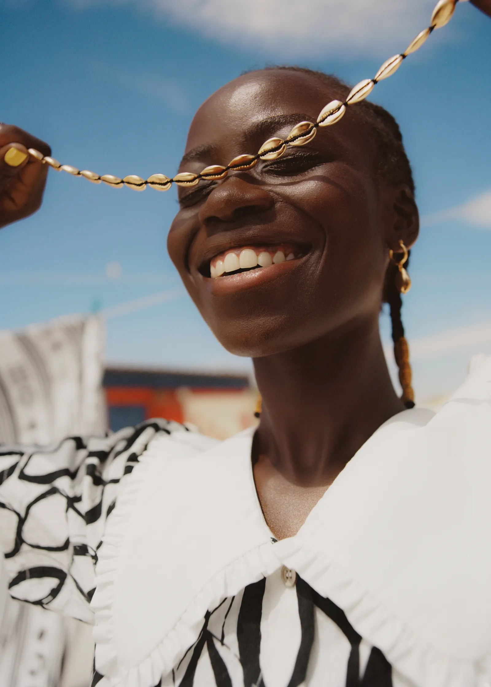 Femme souriante tenant un collier de coquillages devant ses yeux, porte une blouse à col blanc avec motifs noirs, ciel bleu en arrière-plan.