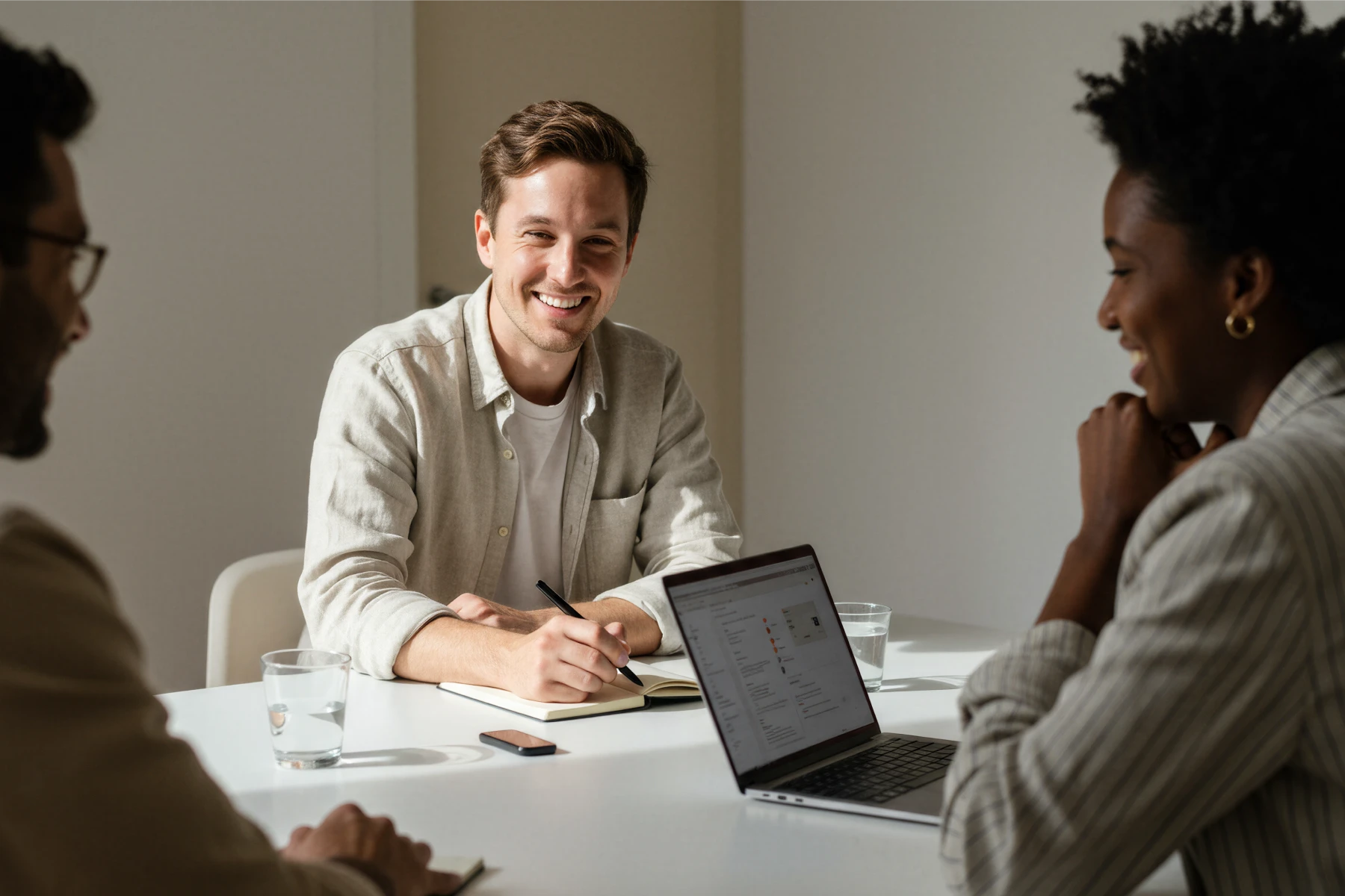Three people smiling and talking around a table, one with a laptop, notebook, and glasses of water.