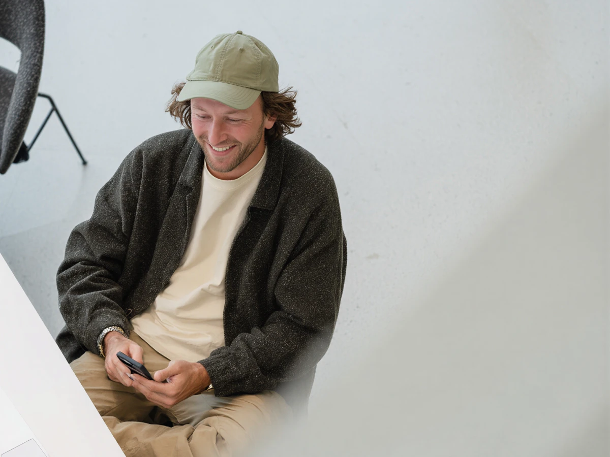 Smiling man in a cap and dark jacket sitting and holding a smartphone looking down.