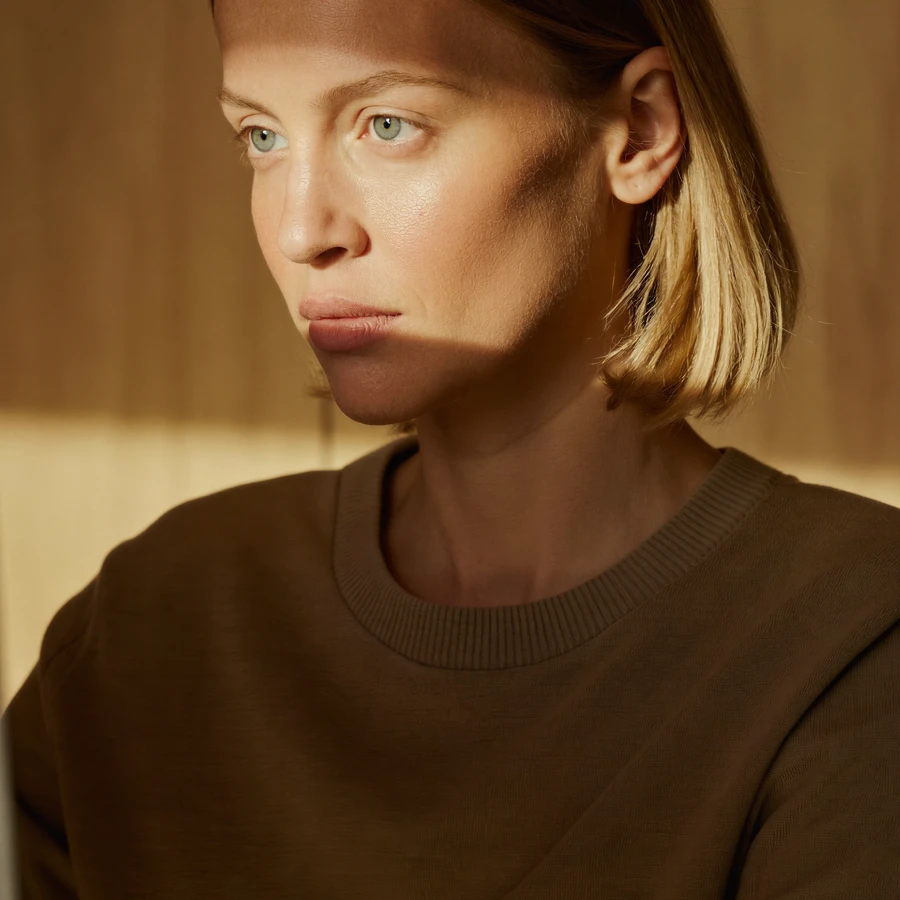 Woman with short blonde hair wearing a brown top, lit by warm sunlight and shadows on her face.