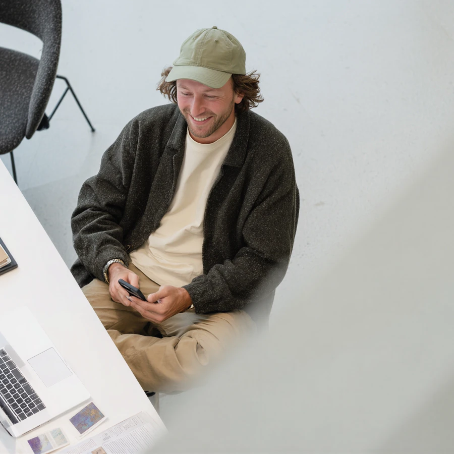 Smiling man in a cap and dark cardigan sitting at desk using a phone with laptop and papers nearby.