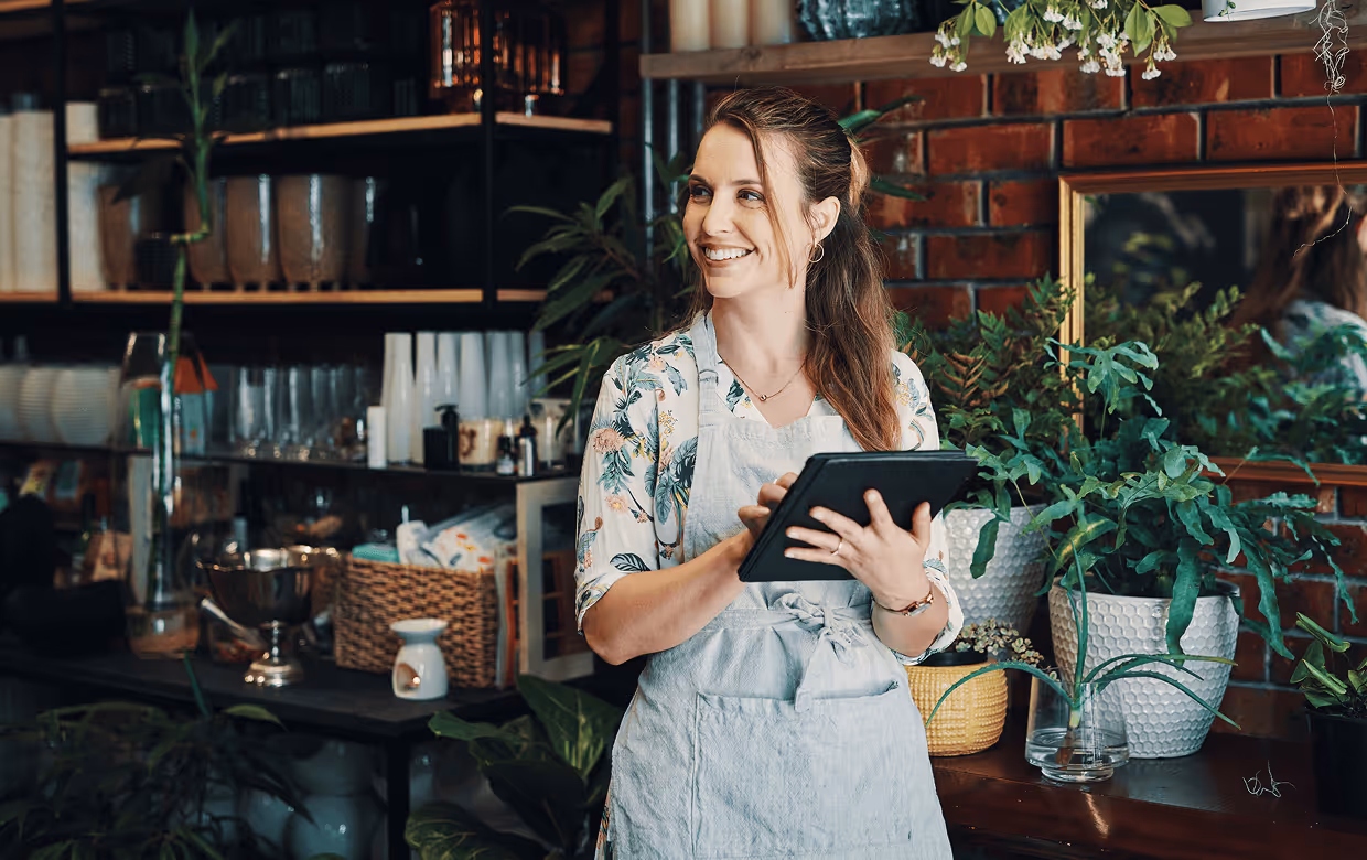 Female small-medium business owner wearing apron holding a tablet and smiling while in her cozy cafe with plants and shelves in the background.