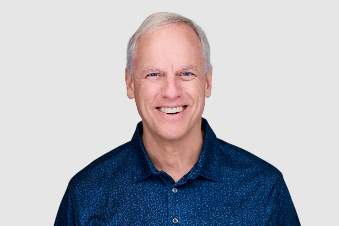 Smiling middle-aged man with gray hair wearing a blue patterned shirt against a light gray background.