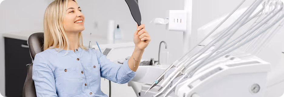 Smiling woman sitting in a dental chair holding a mirror in a modern dental clinic.