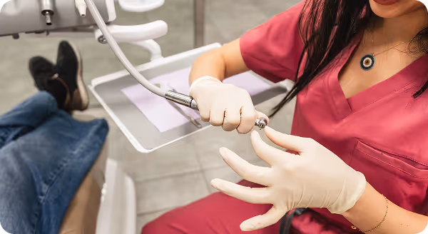 Dental professional in pink scrubs wearing gloves and holding a dental drill near a patient's feet in a dental clinic.