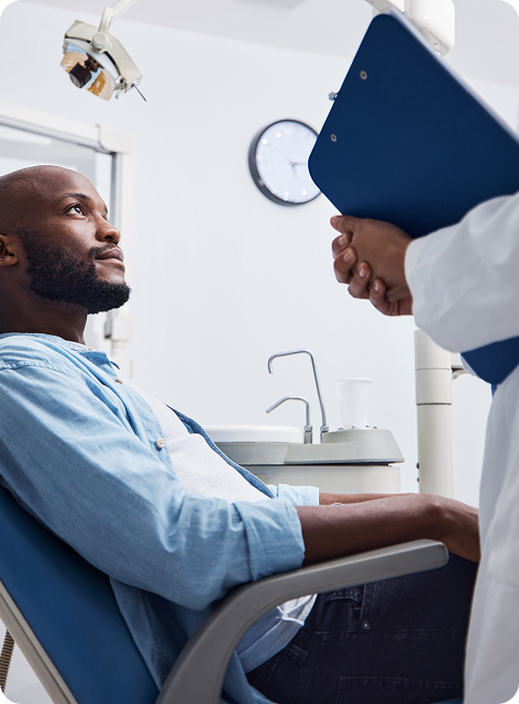Male patient sitting in a dental chair looking attentively at a dentist holding a blue clipboard.