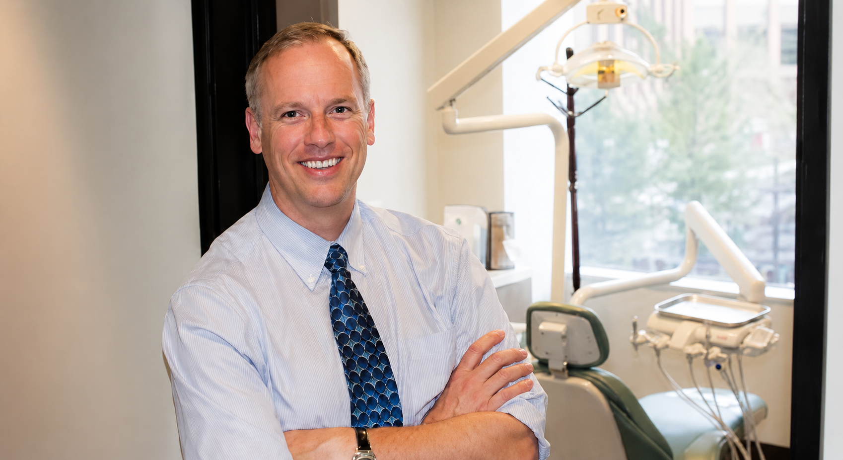 Smiling man in a light blue shirt and patterned tie standing with arms crossed in a dental office.
