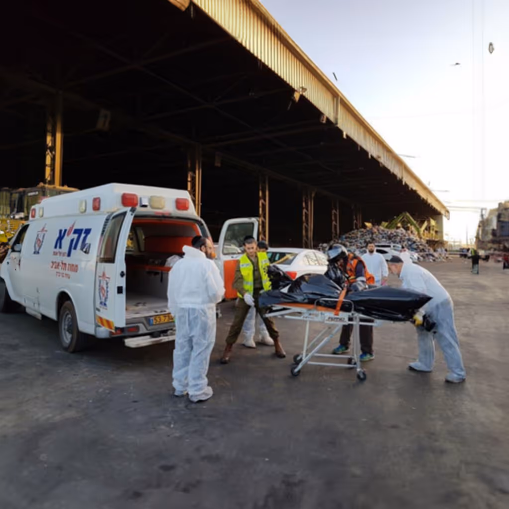 Emergency responders in protective suits loading a body bag on a stretcher into a white ambulance van near a large industrial warehouse.