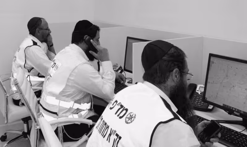 Three men wearing reflective vests and traditional Jewish head coverings working at computers in an office setting.