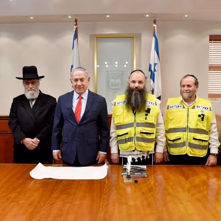 Four men standing behind a wooden table; two Israeli flags are visible in the background, with one man wearing a suit and tie, one in traditional religious attire, and two in yellow emergency response vests.